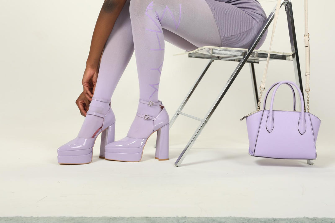 Model seated in chair, leaning forward to display lavender logo tights with ‘MHYK’ lettering and glossy lavender platform MHYK heels, side view against a plain background.