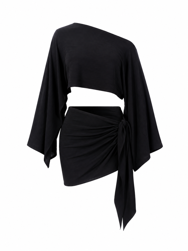 A black asymmetrical one-shoulder top with a draped, cropped silhouette, wide sleeves, and a matching wrapped mini skirt with a side tie, displayed on a white background.